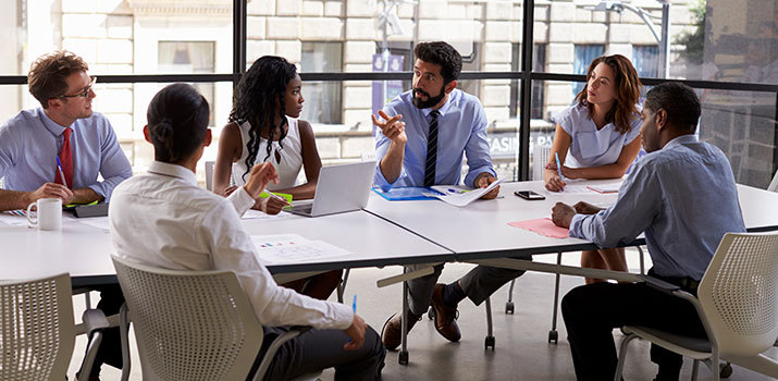 Male in button-down and tie leads a meeting with a mixed group of colleagues at a large conference table with windows in the background.