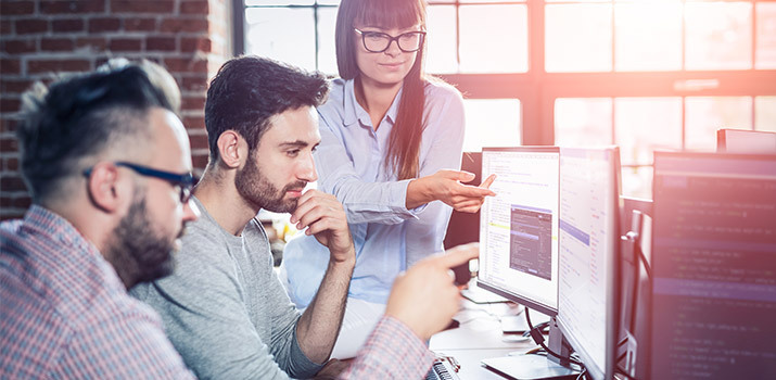 Coworkers at a software development office discuss a project while pointing at computer screens.