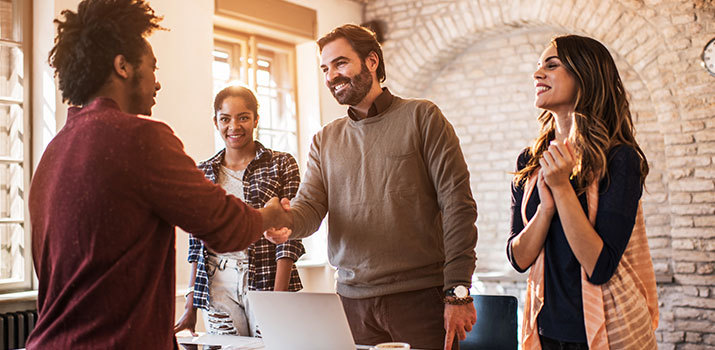 Tech startup employees in business casual are welcoming a newly hired team member with a hand shake.